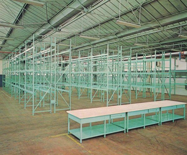 Uno spazio industriale adibito a magazzino, caratterizzato da file di scaffalature metalliche vuote di colore verde chiaro e da un lungo banco da lavoro in primo piano.