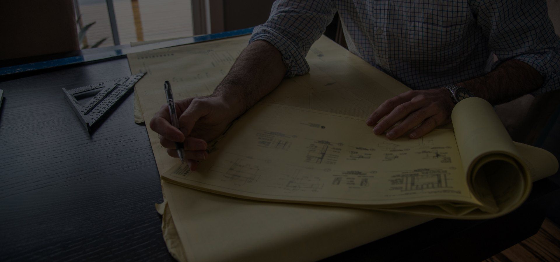 Person drawing on architectural plans. A rolled-up paper is on a desk. A pencil is used on the drawings.
