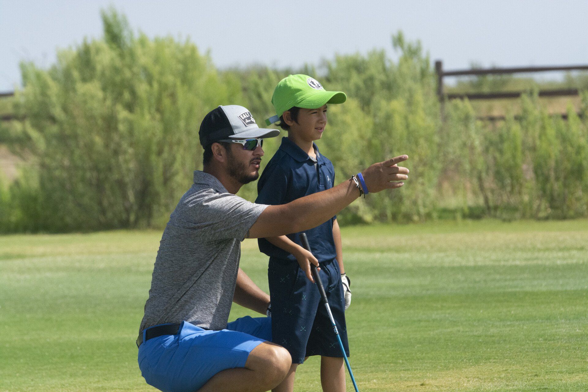 Two golfers on a green, one kneeling and pointing while holding a club, with trees in the background