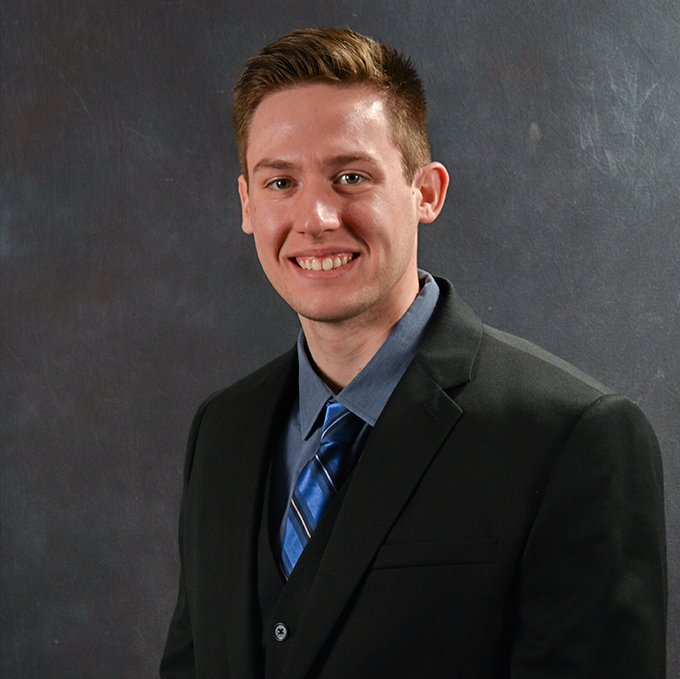 Studio portrait of a smiling man in a dark suit and blue tie against a gray backdrop