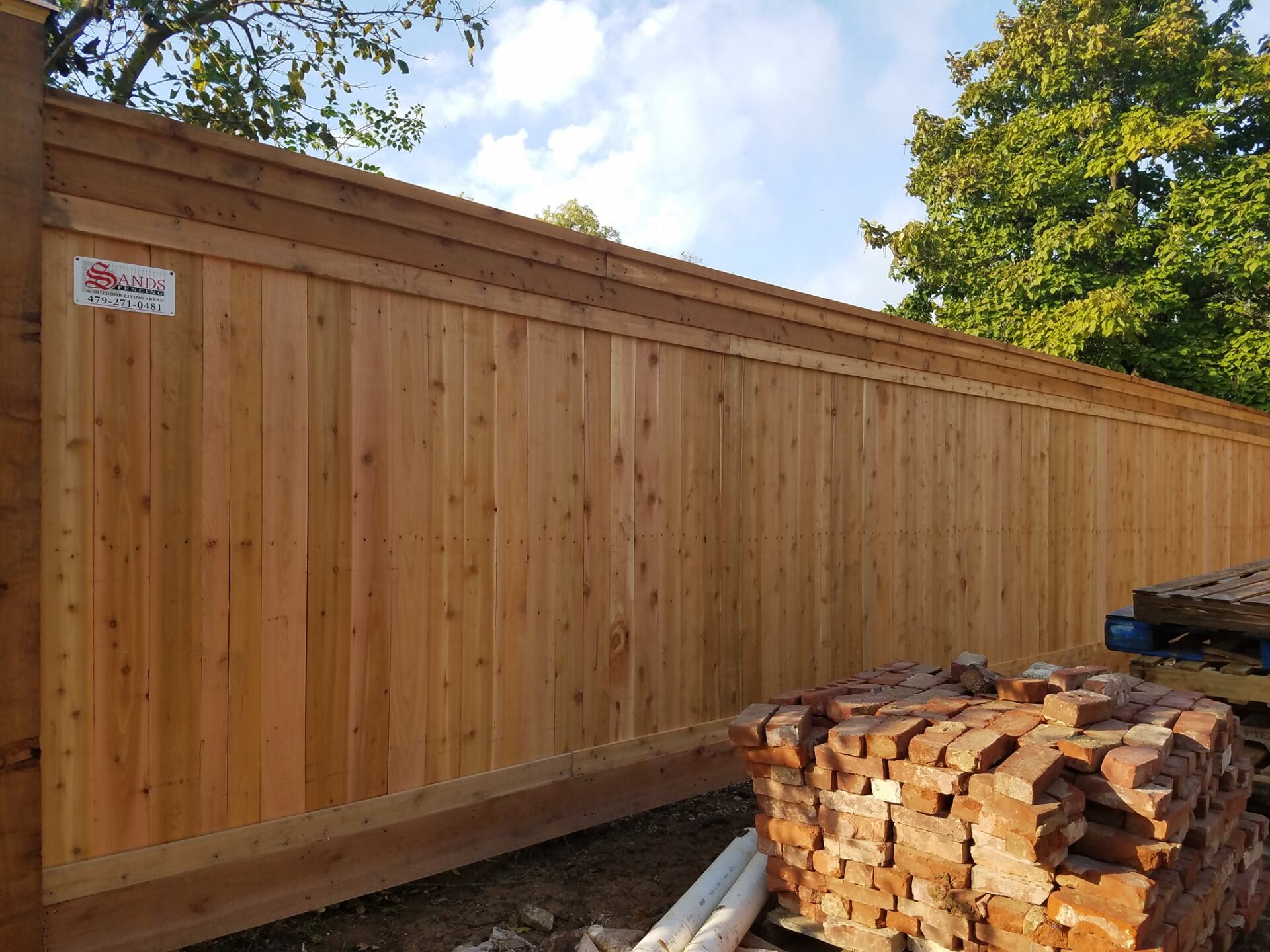 Wooden privacy fence along a yard with a stack of split firewood in the foreground under a blue sky.