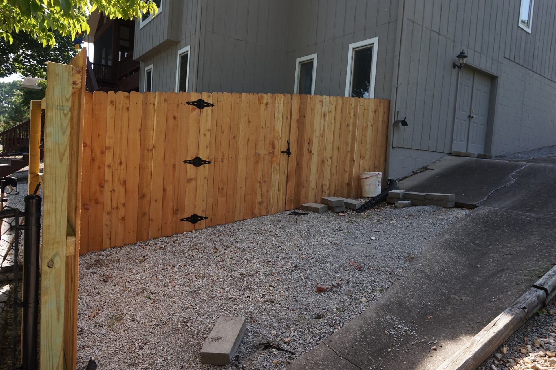 Wooden fence beside a gray house with a gravel driveway and sloped asphalt path