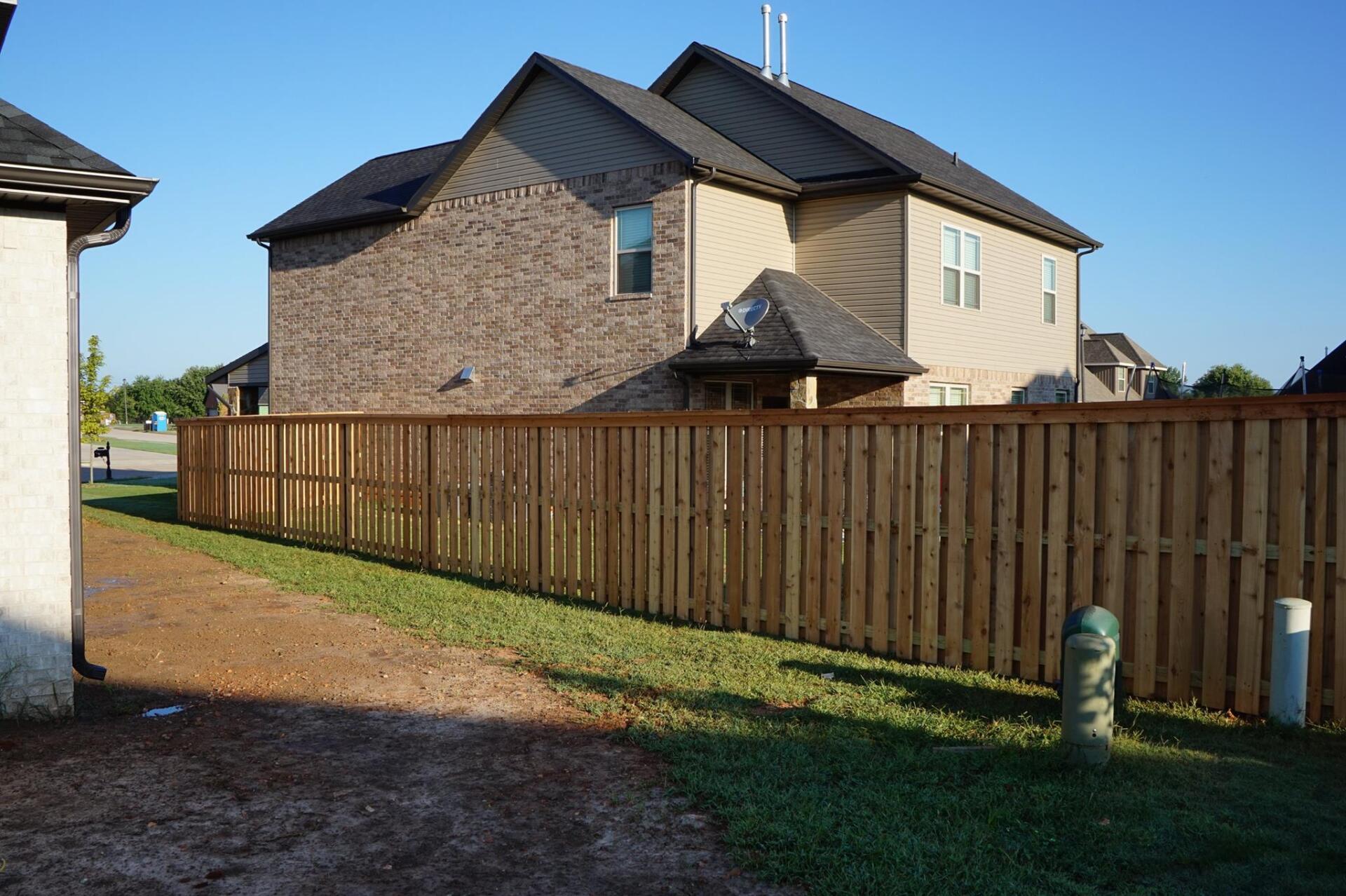 Wooden fence along a suburban backyard beside a brick house and gravel driveway under a clear sky