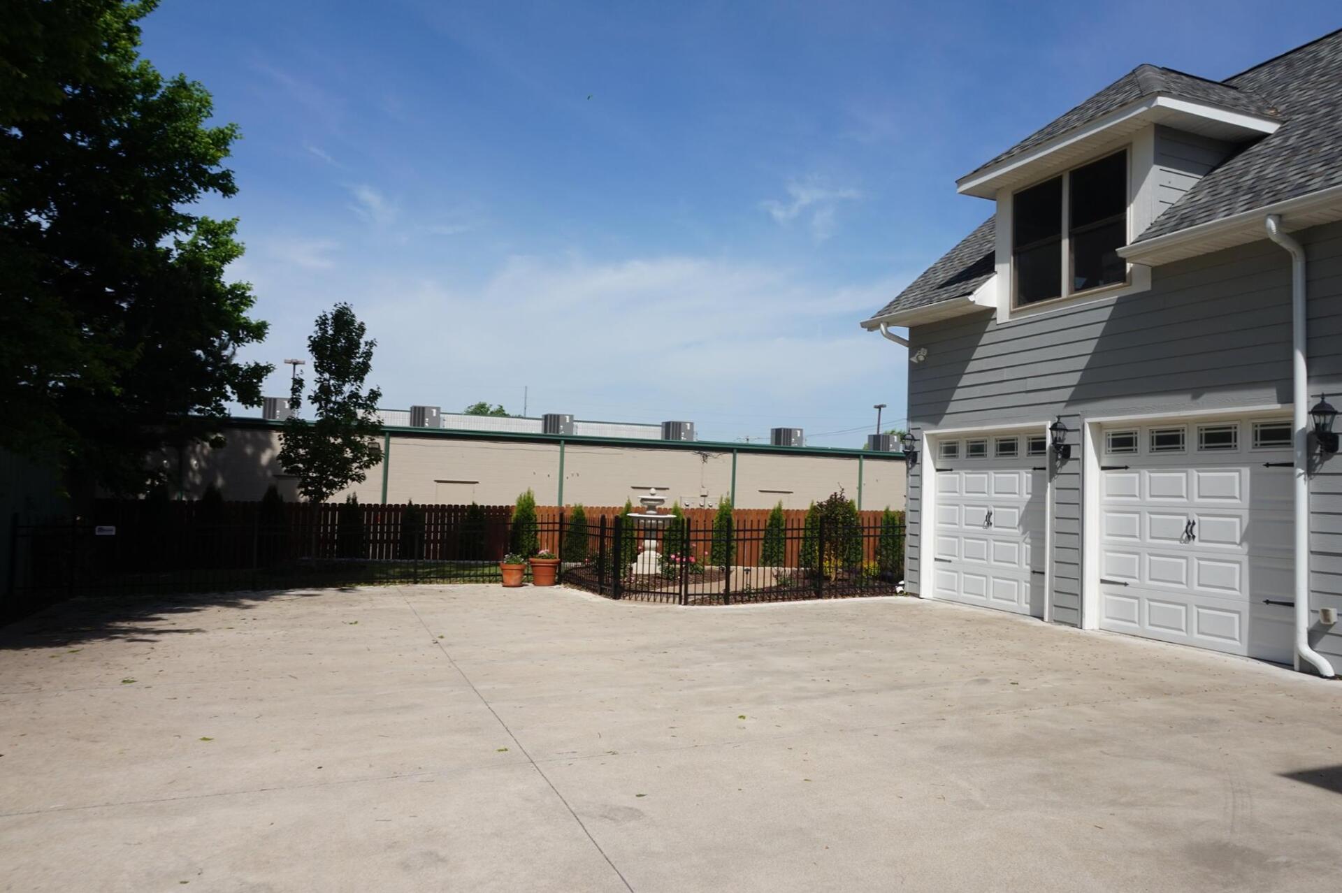 Empty concrete parking area beside a gray building with garage doors and a small fenced garden under a blue sky
