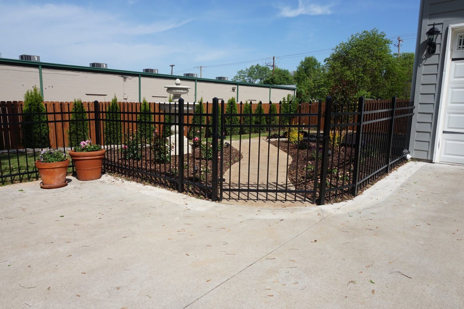 Black metal gate around a small landscaped patio beside a light-gray house on a sunny day
