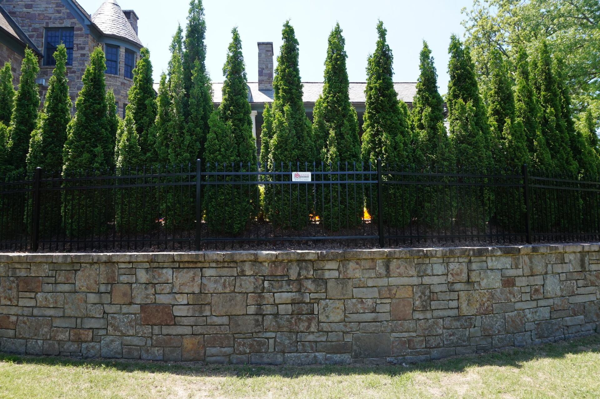Stone wall with tall green hedges and a black fence in front of a house