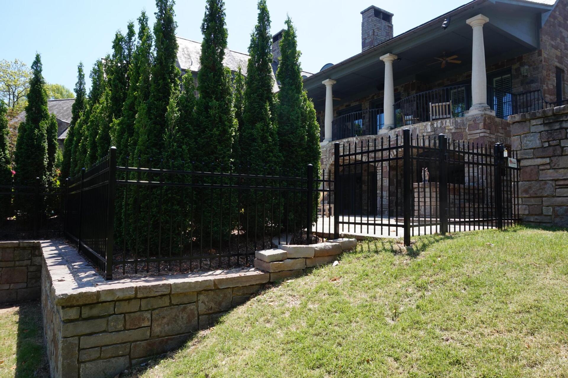 Stone house with porch behind black fence, tall evergreens, and grassy sloped yard