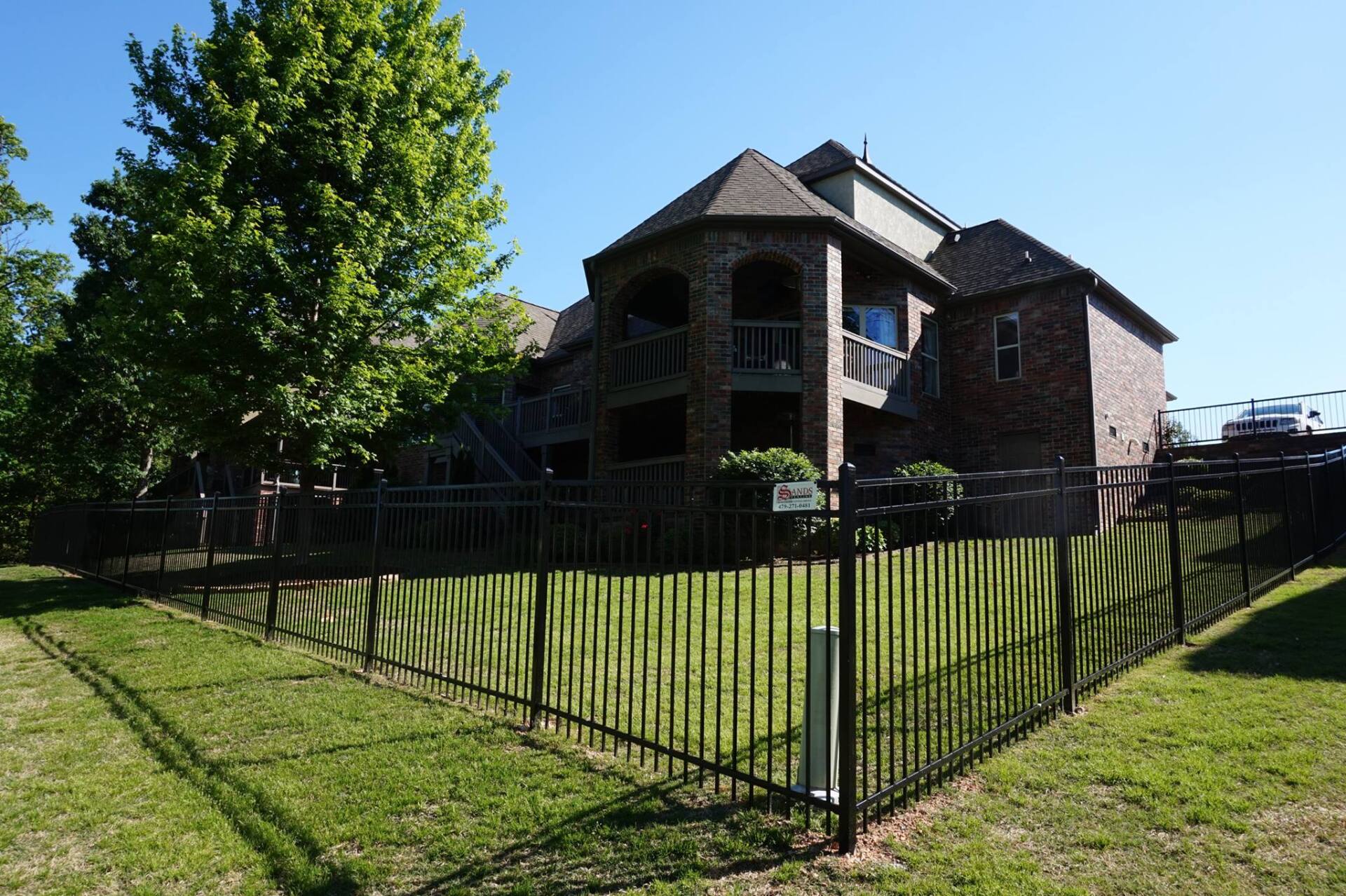 Brick house with a steep roof behind a black fence, beside a large green tree on a sunny day