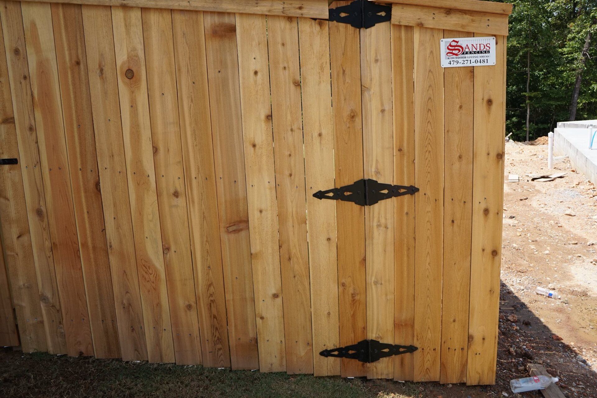 Wooden fence gate with black hinges and a small sign, beside a dirt path outdoors