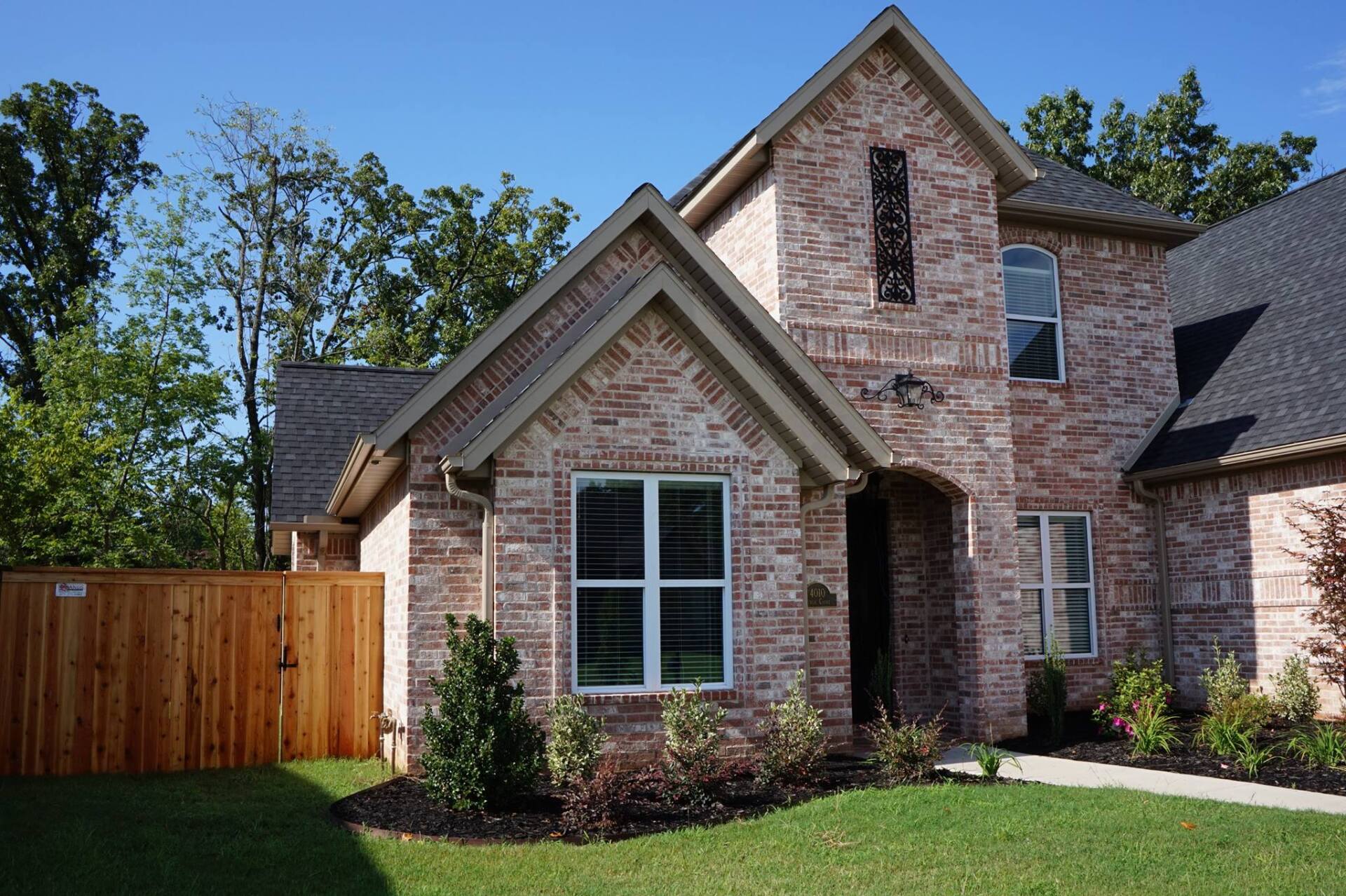Brick suburban house with stone facade, wooden fence, and green lawn under a clear blue sky
