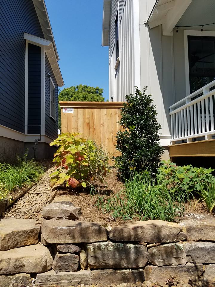 Narrow landscaped side yard with a small tree, shrub, and stone retaining wall between two houses