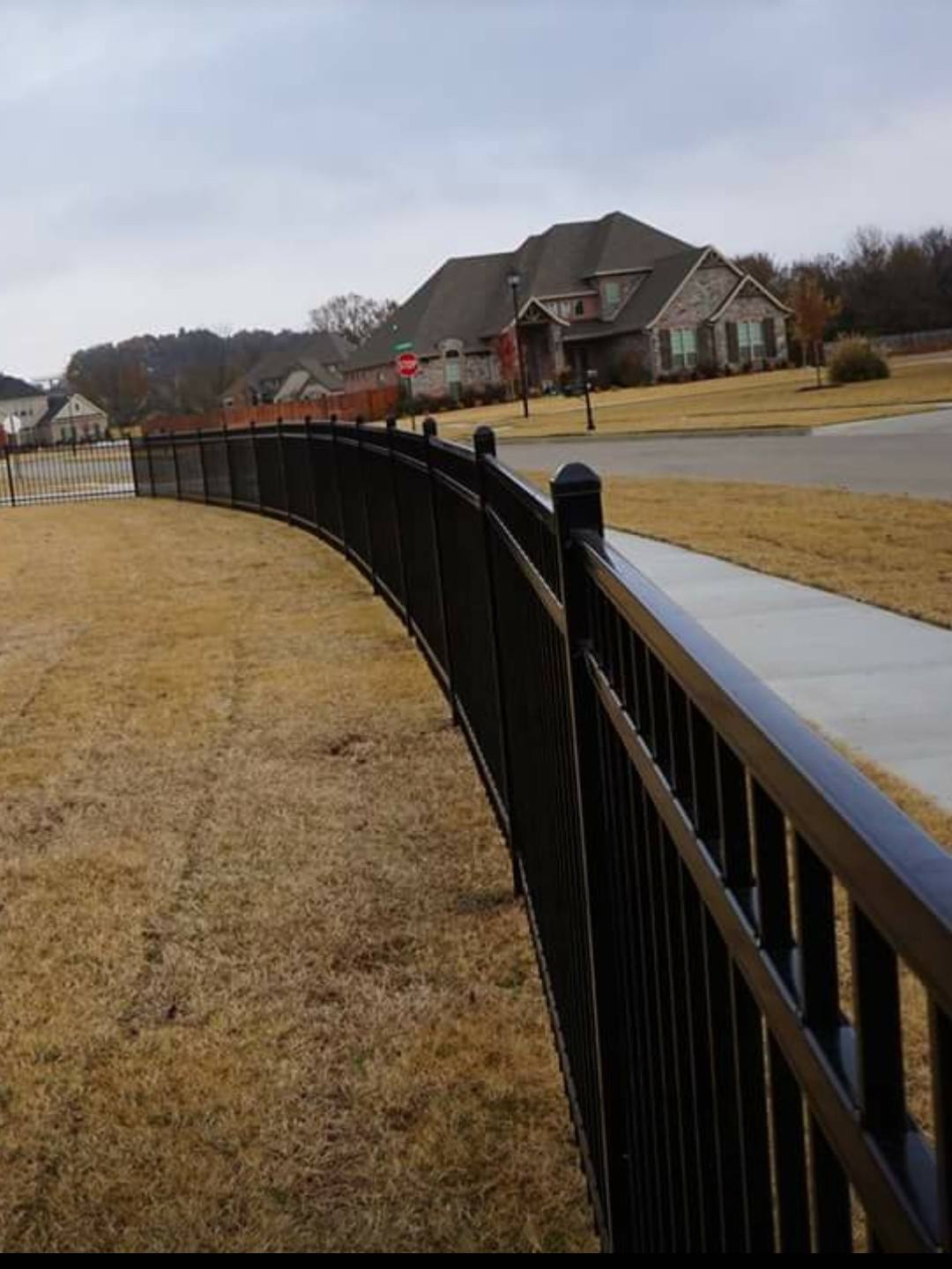 Curved black fence along a brown lawn beside a sidewalk, with houses in the distance on a cloudy day