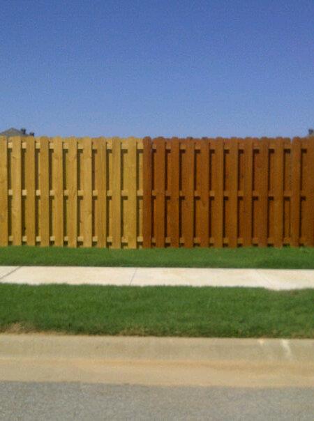 Brown wooden fence beside a green lawn, sidewalk, and road under a clear blue sky