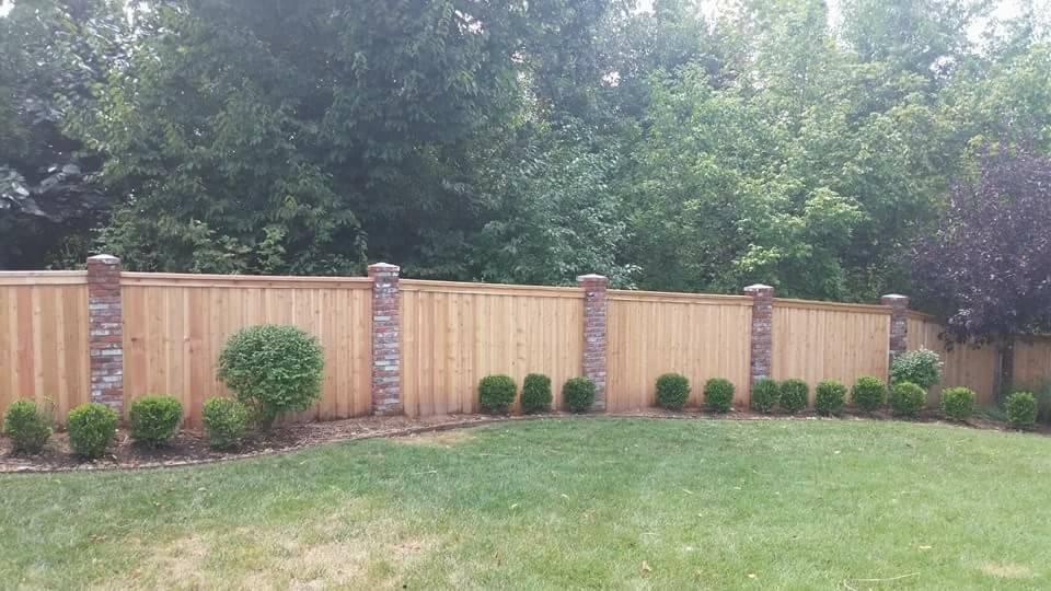 Wooden privacy fence with brick posts and trimmed shrubs along a grassy backyard edge.