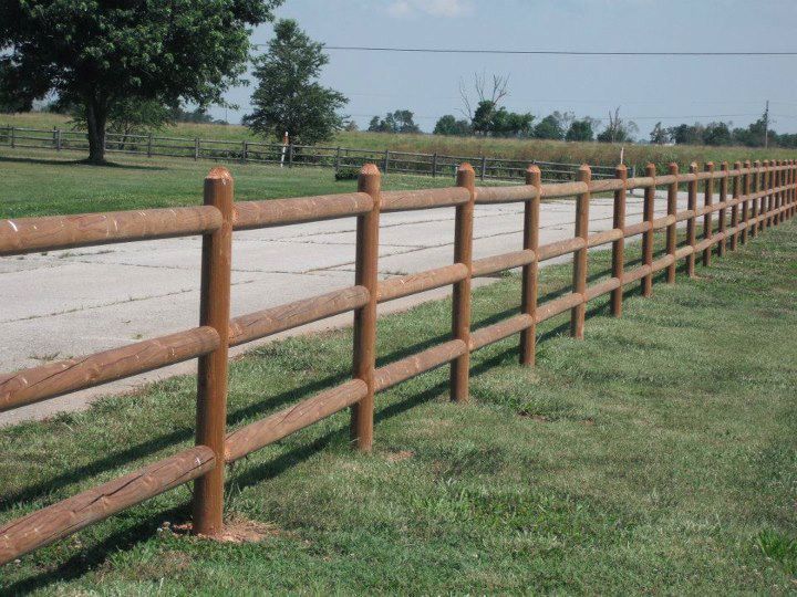 Brown wooden fence running along a dirt road beside green grass in a rural field.