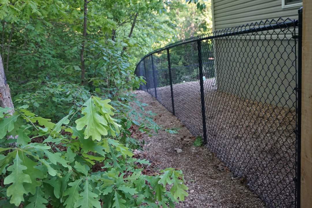 Chain-link fence along a wooded yard beside a beige house, with gravel and leafy greenery.