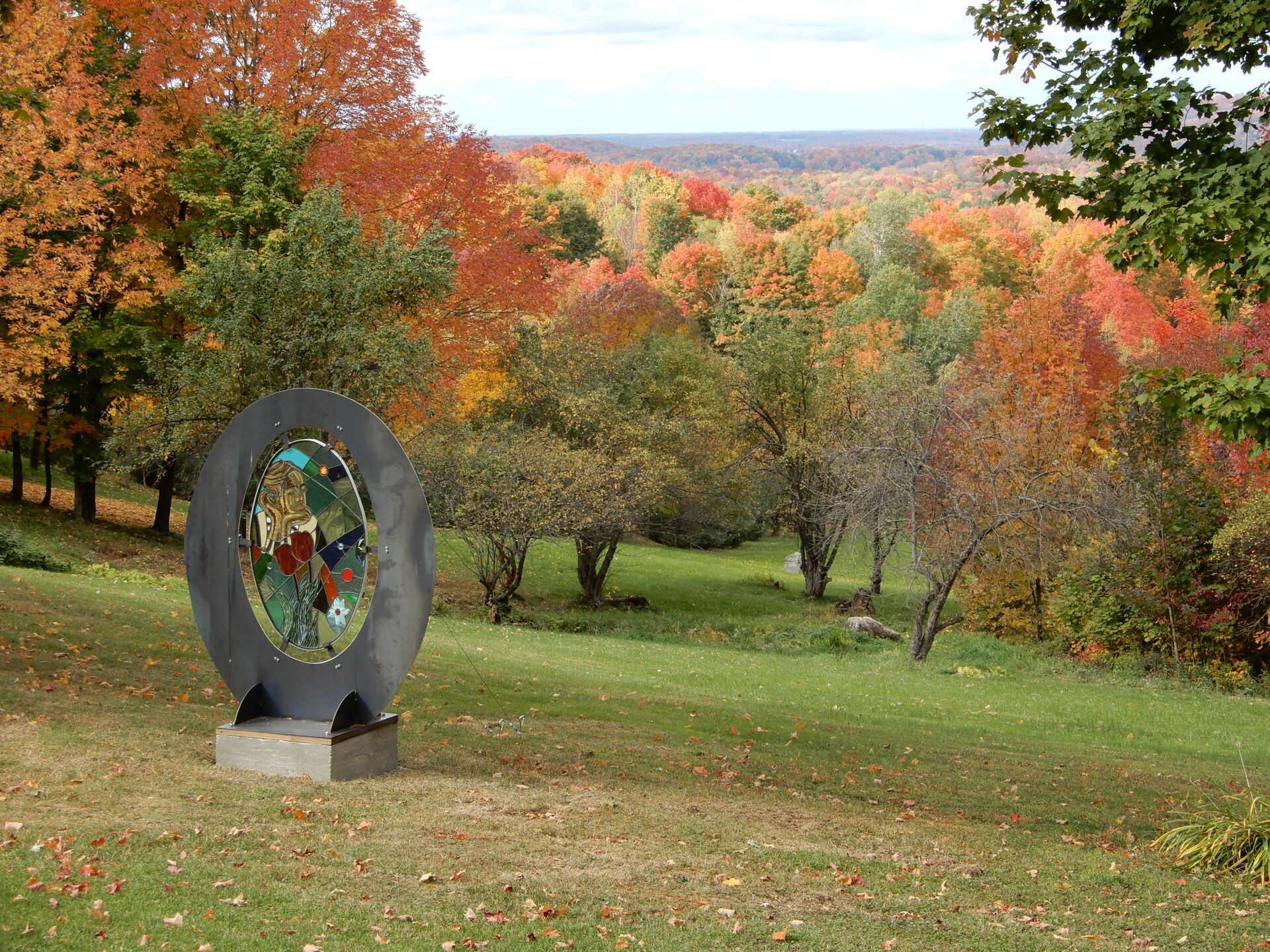 Sculpture ovale sur une colline verdoyante, entourée d'arbres d'automne vibrants, surplombant une forêt lointaine.