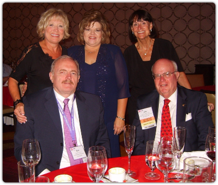 Five people at a table; three women standing behind two men. They are smiling. Formal event.