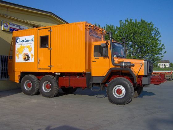 Camion arancione da spedizione con cabina personalizzata, parcheggiato vicino a un edificio.