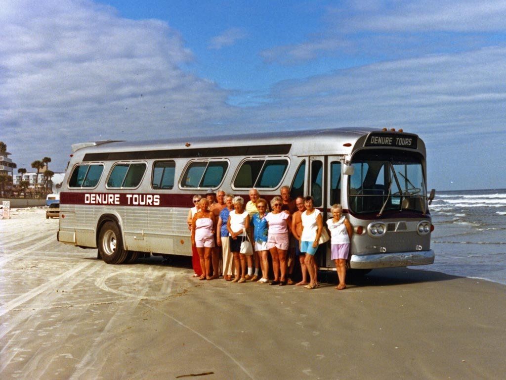 People stand near a vintage silver bus on a beach under a blue sky. The bus reads 
