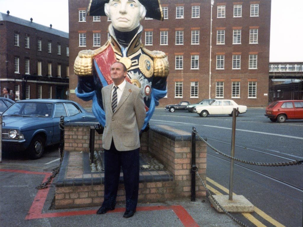 Man standing in front of a large statue of a naval officer. Cars and brick building in the background.