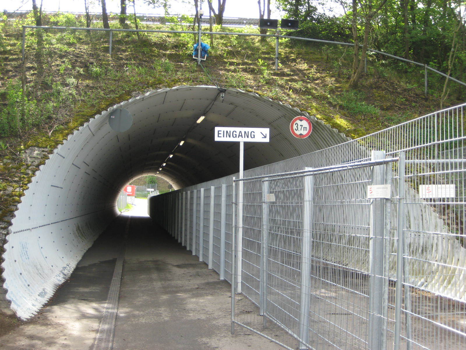 Tunneleingang mit umzäuntem Fußweg und Straßenschild unter einem grasbewachsenen Damm