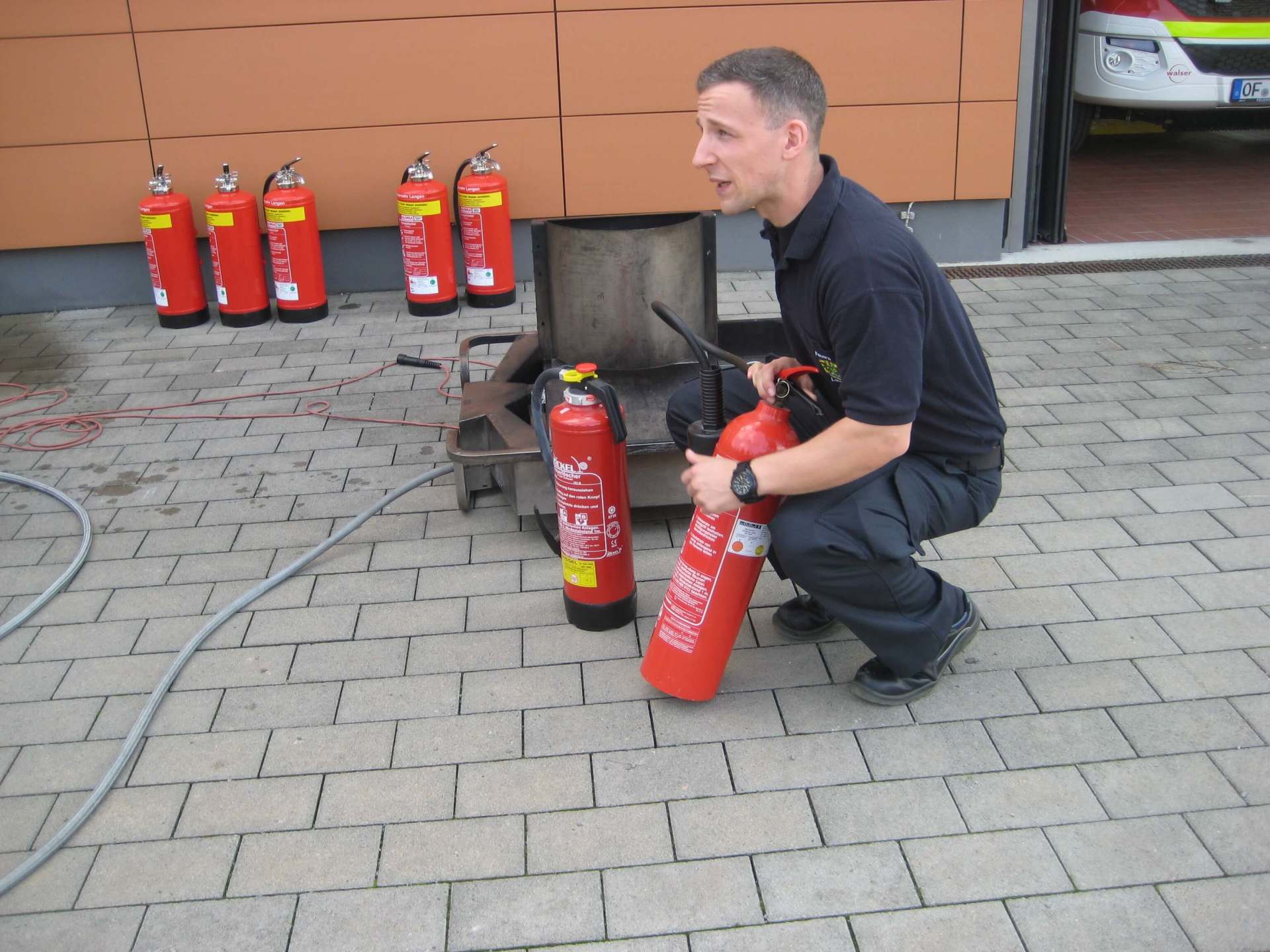 Ein Mann kniet neben mehreren roten Feuerlöschern auf einem gepflasterten Untergrund und überprüft einen davon in der Nähe eines offenen Koffers.