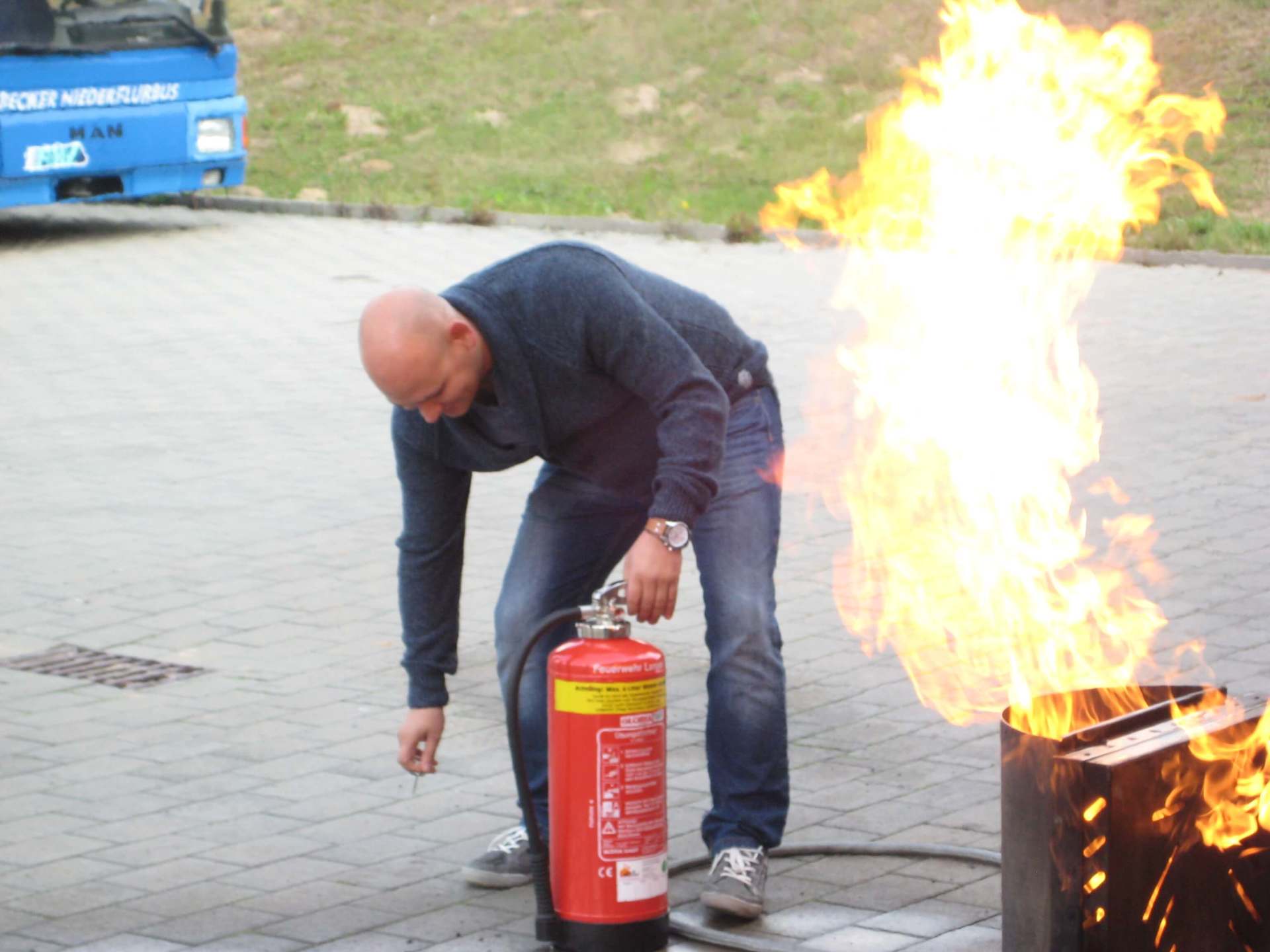Eine Person beugt sich über eine große Flamme auf einer gepflasterten Fläche und hält einen Feuerlöscher in der Hand.