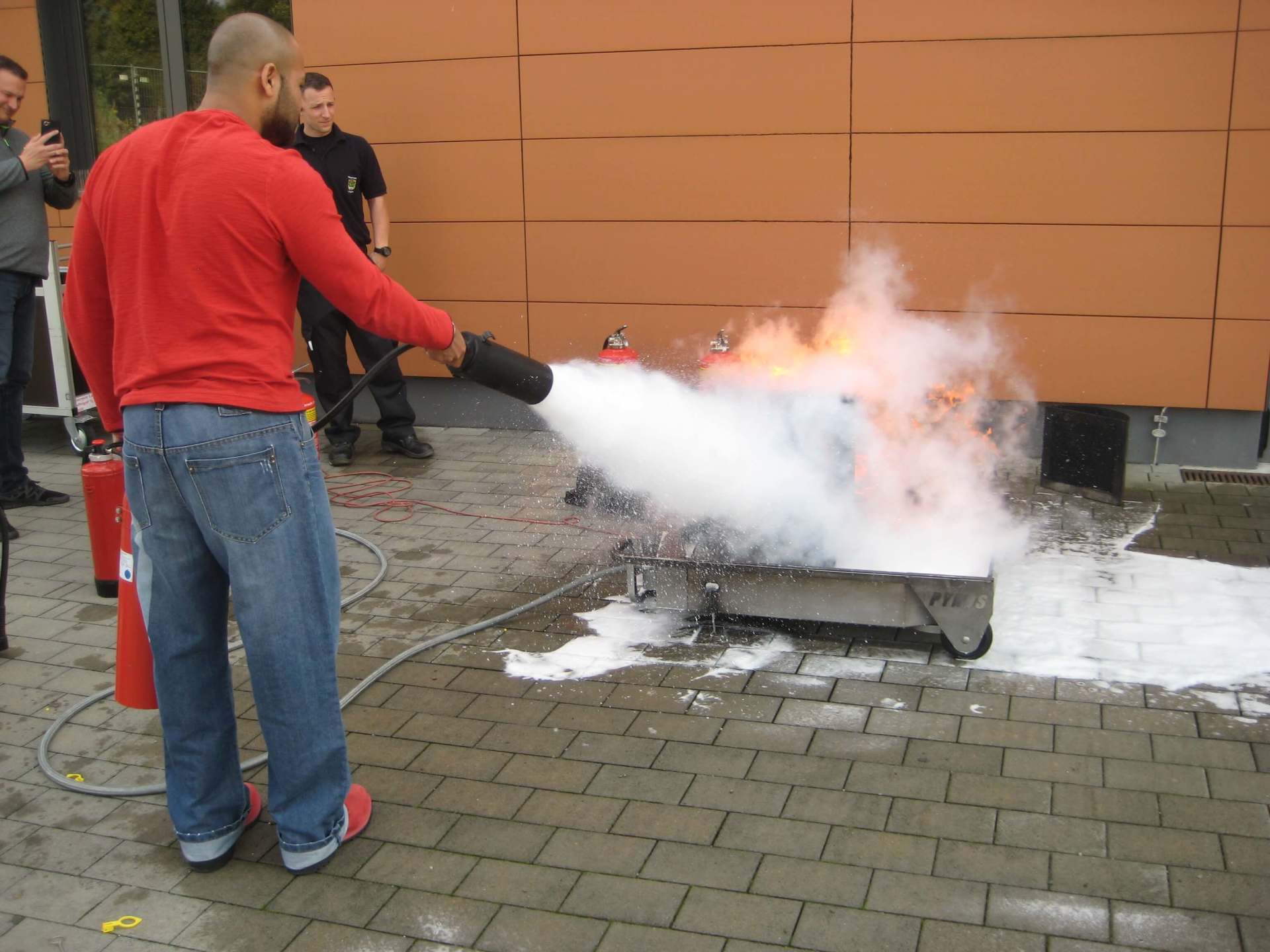 Eine Person benutzt einen Feuerlöscher, um ein kleines Feuer im Freien in der Nähe einer Backsteinmauer zu löschen.
