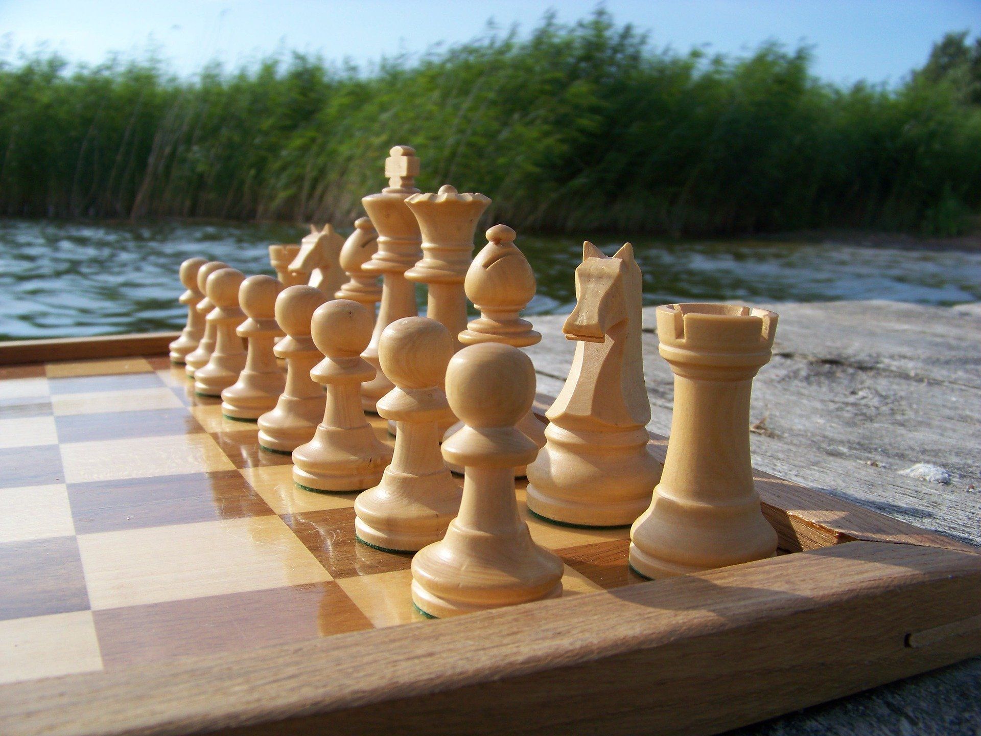 Ein hölzernes Schachspiel auf einem Brett im Freien am Wasser, im Hintergrund verschwommenes Grün.