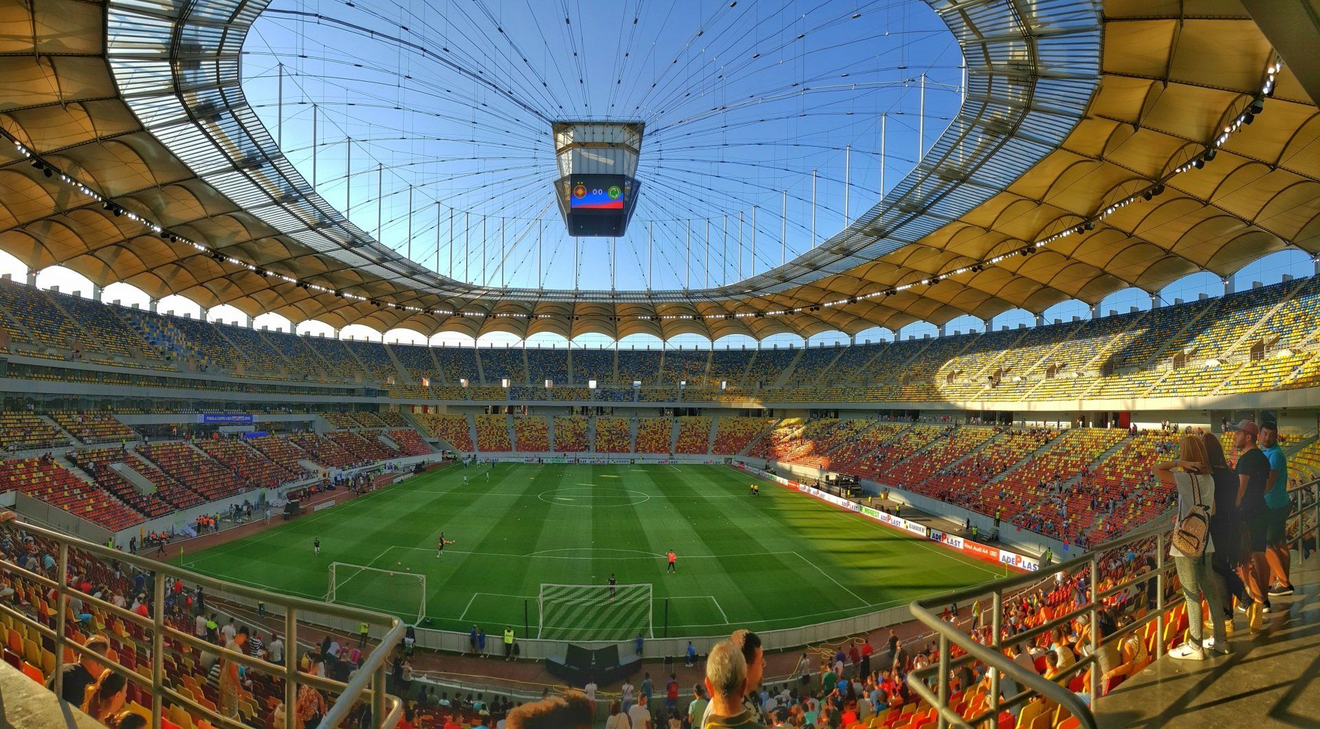 Weitläufiges Fußballstadion mit grünem Rasen, vollbesetzten Tribünen und einem großen runden Dach unter blauem Himmel
