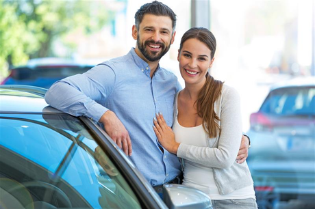 Couple smiling, arm in arm, leaning on car in showroom.