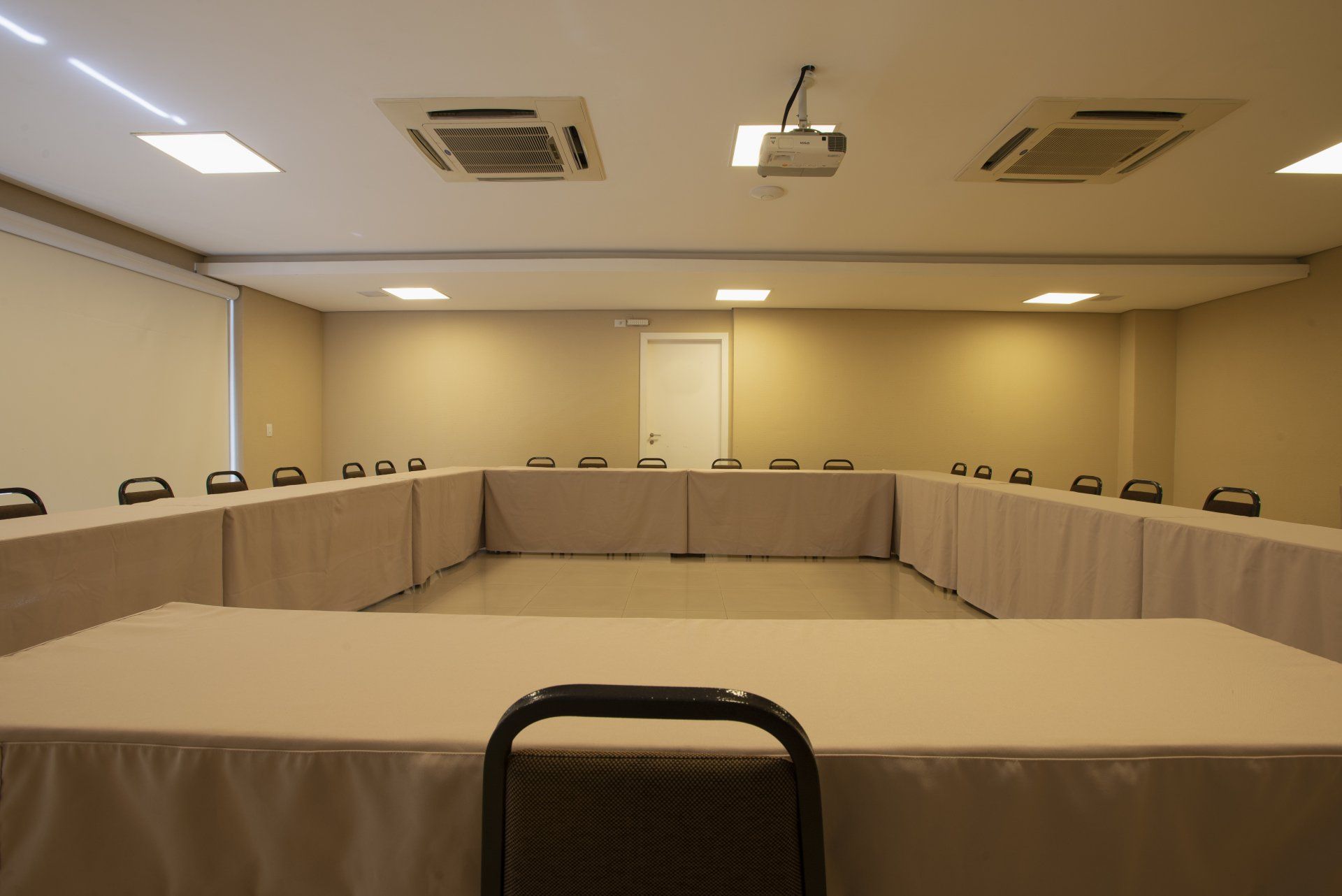 Sala de conferências vazia com paredes bege e uma mesa em formato de U coberta com uma toalha de mesa clara.