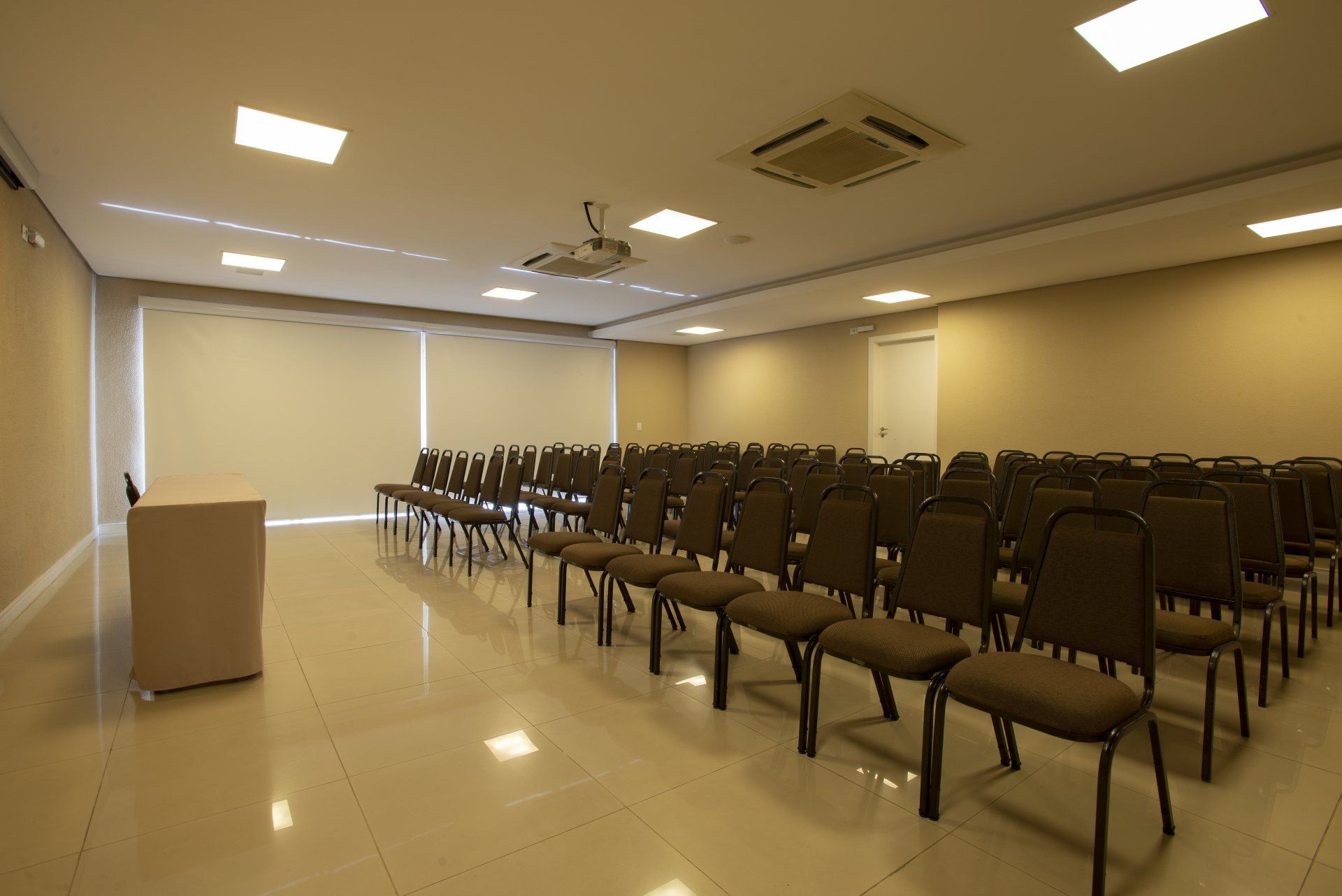 Sala de conferências vazia com fileiras de cadeiras, tela de projeção e pódio. Paredes bege, teto branco e piso brilhante.