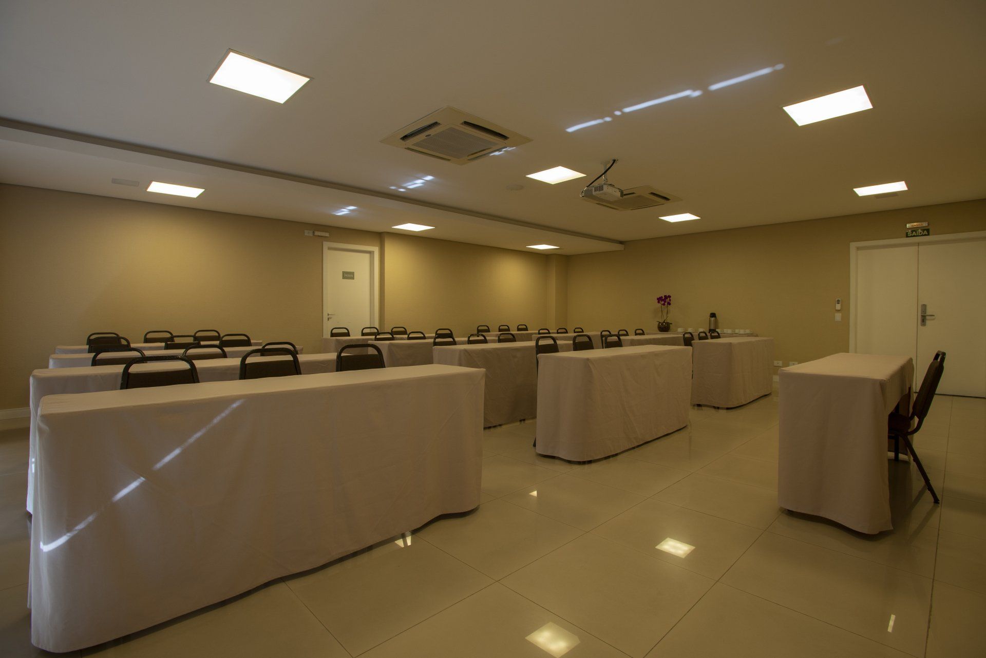 Sala de conferências com fileiras de mesas cobertas com tecido bege e cadeiras pretas.