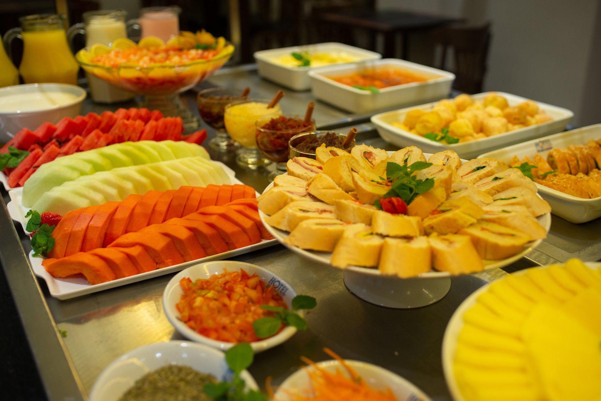 Mesa de bufê com frutas fatiadas, pães, molhos e outros alimentos para café da manhã.