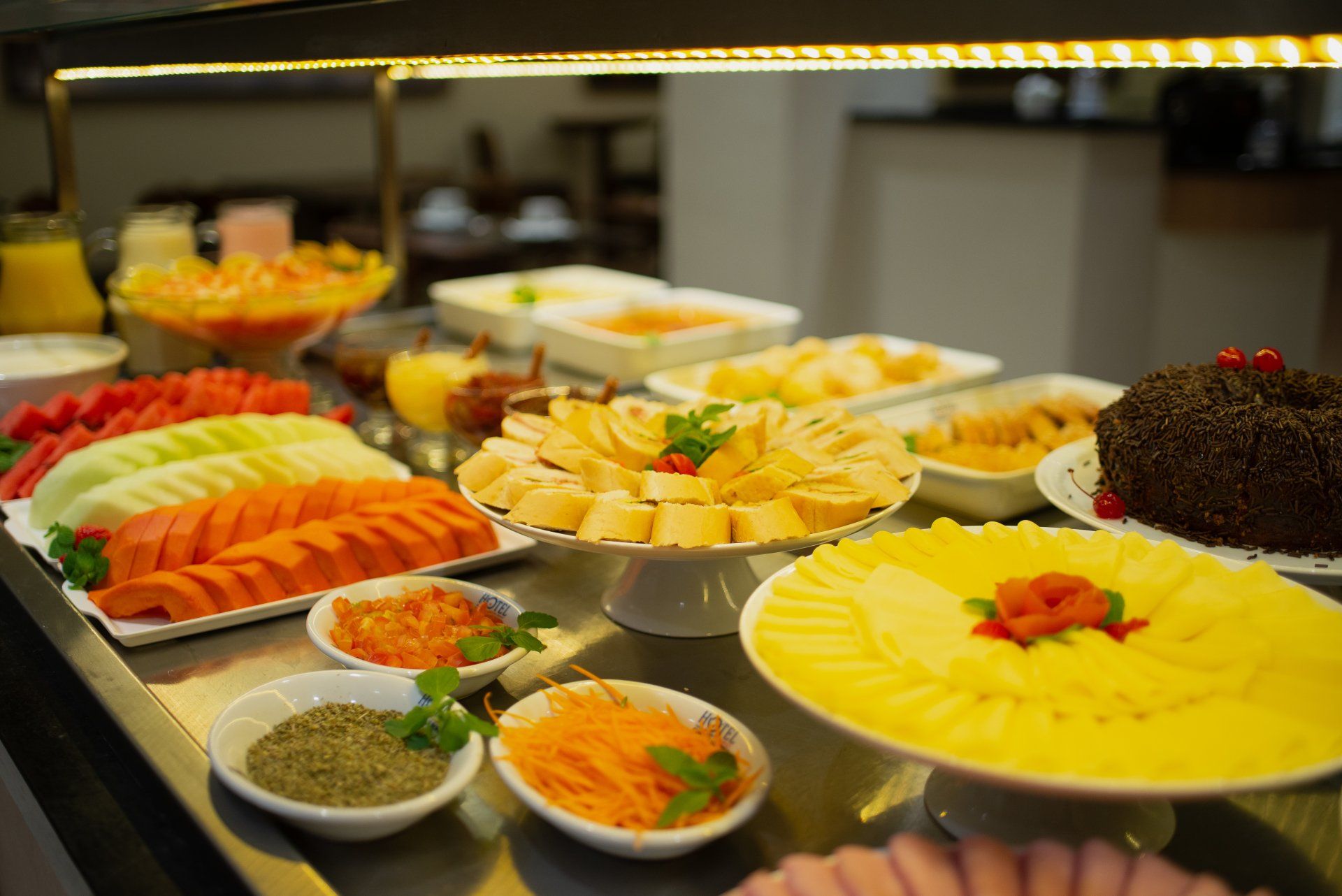Buffet de café da manhã com frutas fatiadas, queijos, pães e bolos.