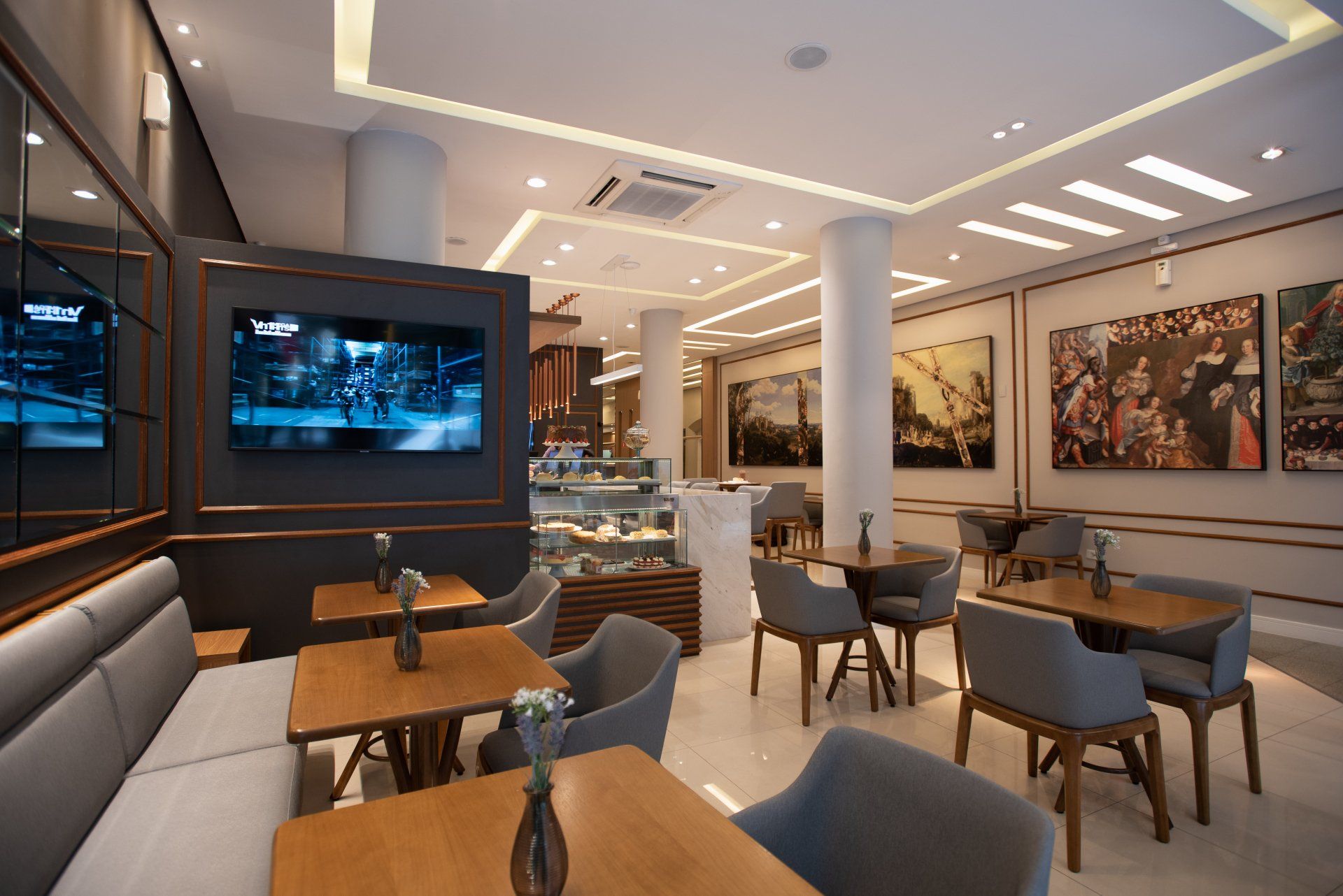 Interior de um café com mesas, cadeiras, arte nas paredes e uma tela de televisão.