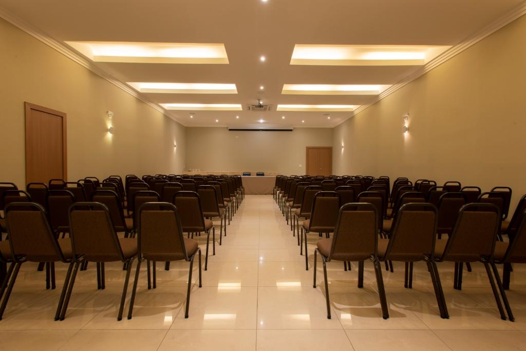 Sala de conferências vazia com fileiras de cadeiras marrons, paredes creme e uma tela de projeção.