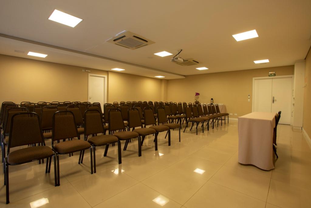 Sala de conferências vazia, bege, com fileiras de cadeiras, projetor e um pódio.