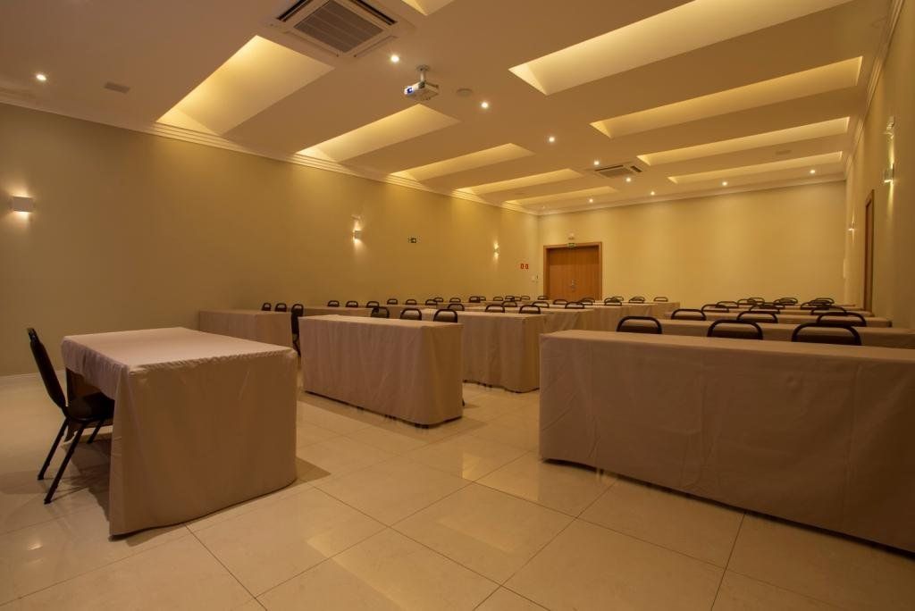 Sala de conferências com mesas cobertas com toalhas bege, cadeiras e iluminação embutida.