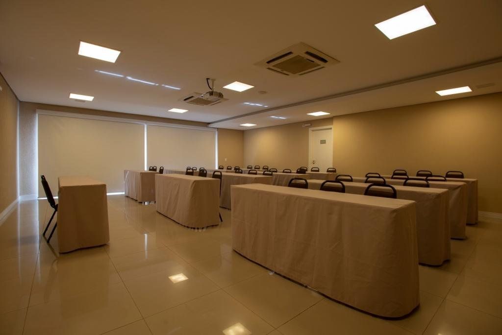 Sala de aula vazia, bege, com mesas e cadeiras cobertas, um projetor e iluminação embutida.