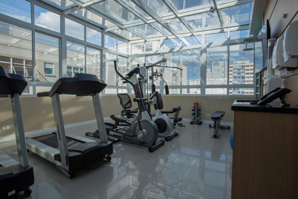 Academia com esteiras, elíptico e aparelho de musculação; um espaço iluminado, cheio de janelas e com vista para a cidade.