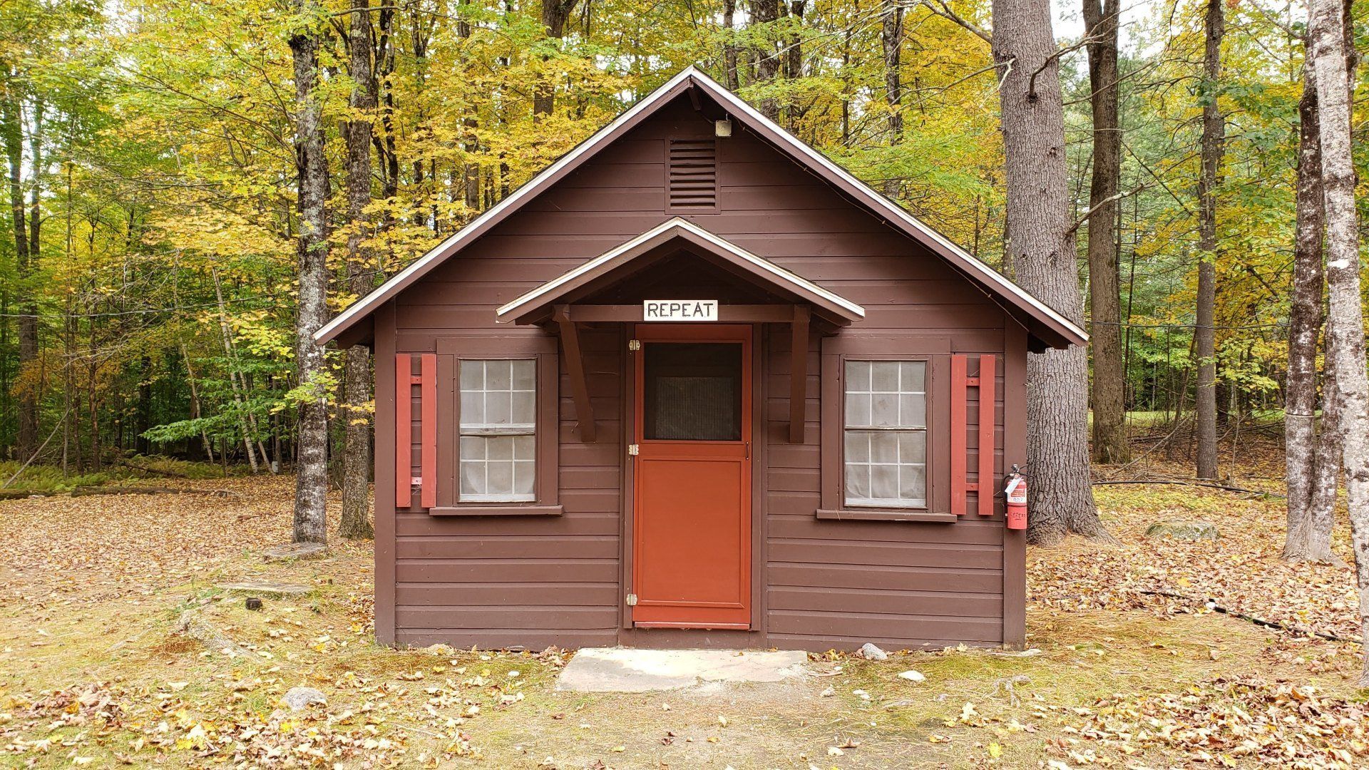 A small brown house with a red door is in the middle of a forest.