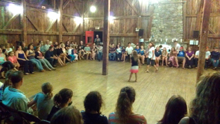 A group of people are sitting in a circle watching a girl do a handstand on a wooden floor.