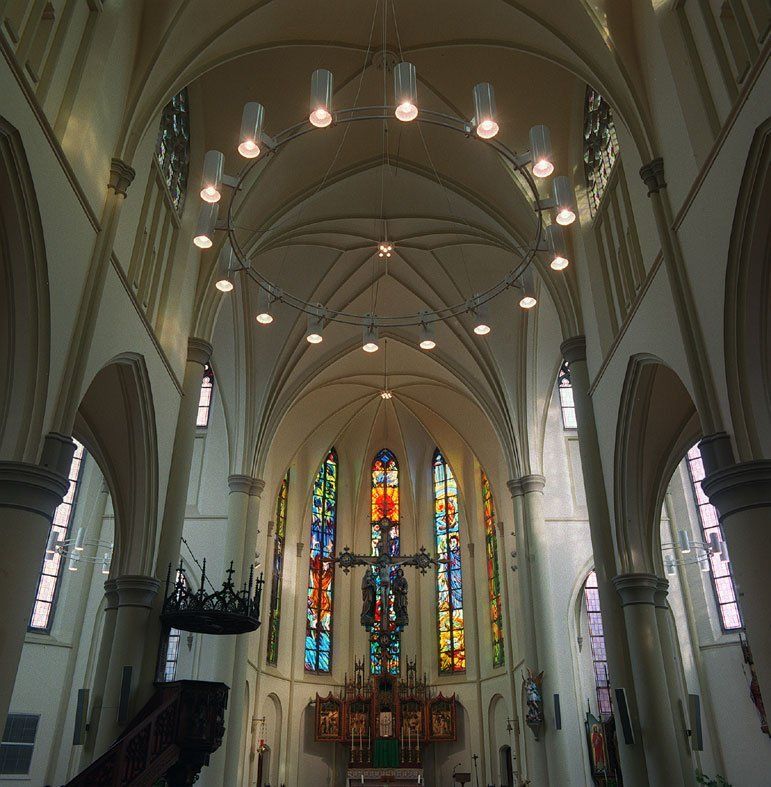 Innenansicht einer Kathedrale mit Gewölbedecken, Hängelampen und farbenfrohen Buntglasfenstern am Altar