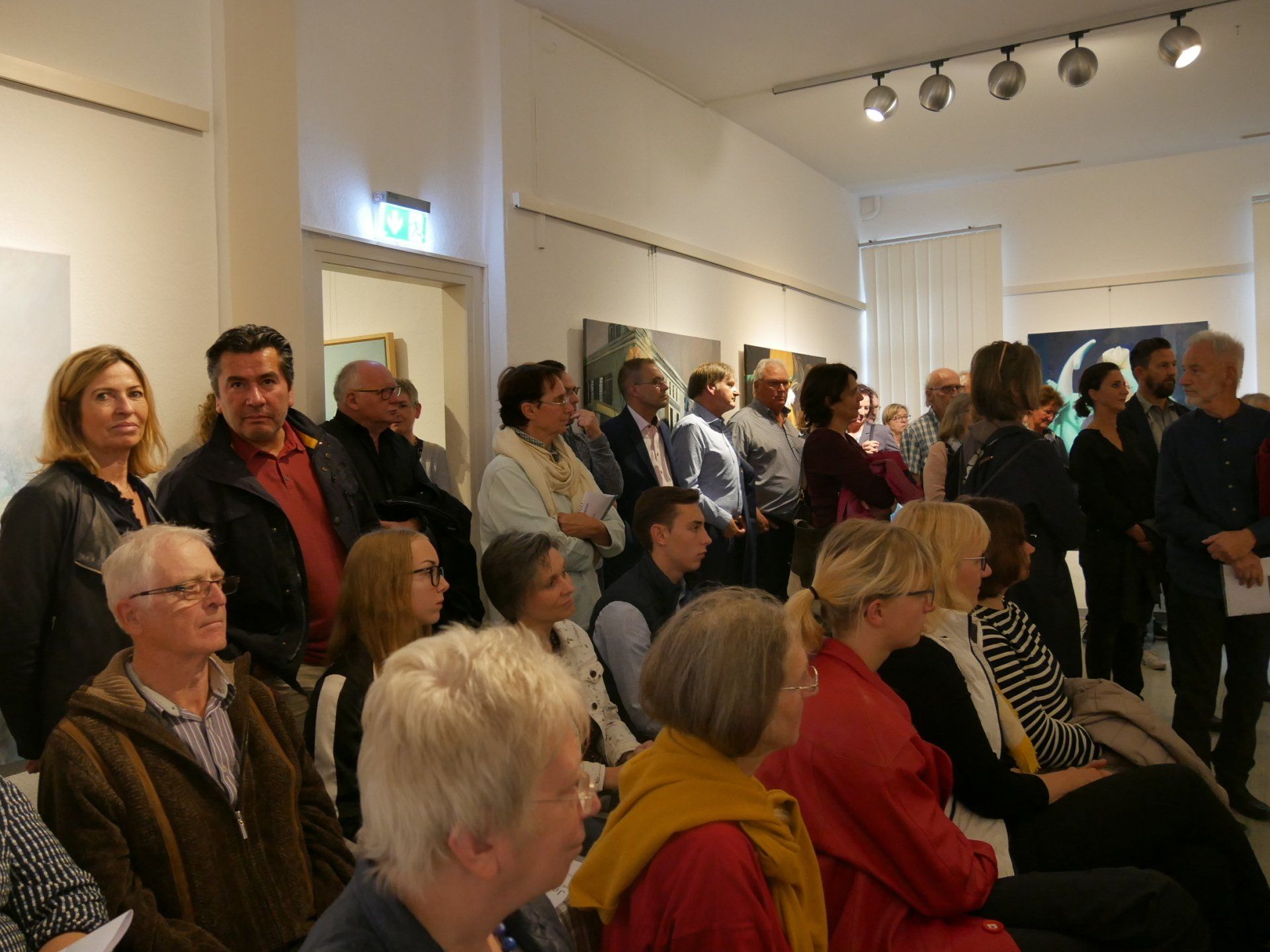 Zuschauer sitzen auf einer Galerie und verfolgen eine Veranstaltung in einem hellen, weißen Raum.
