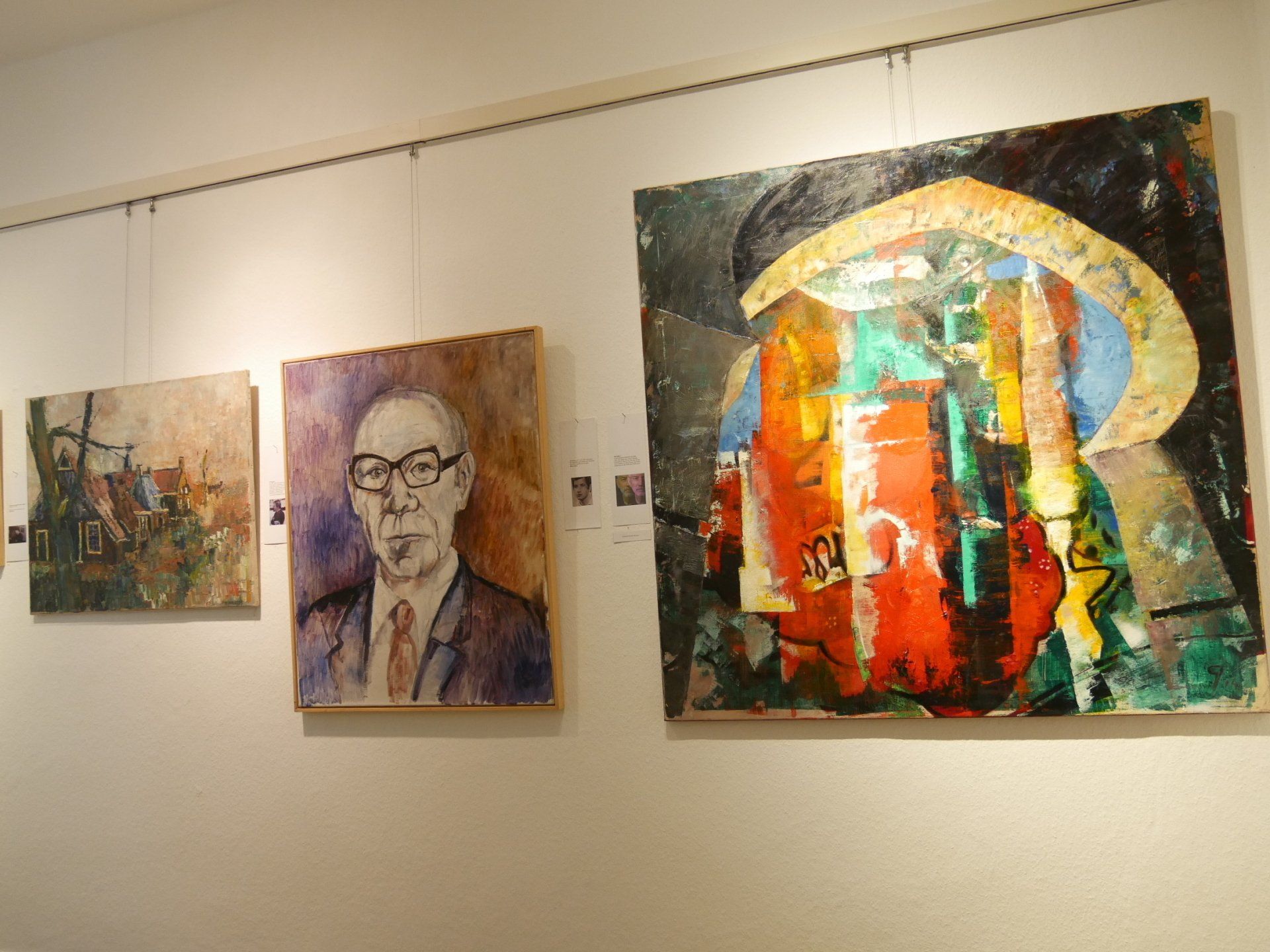 Drei farbenfrohe Gemälde, darunter ein Porträt und abstrakte Szenen, hängen an einer Galeriewand.