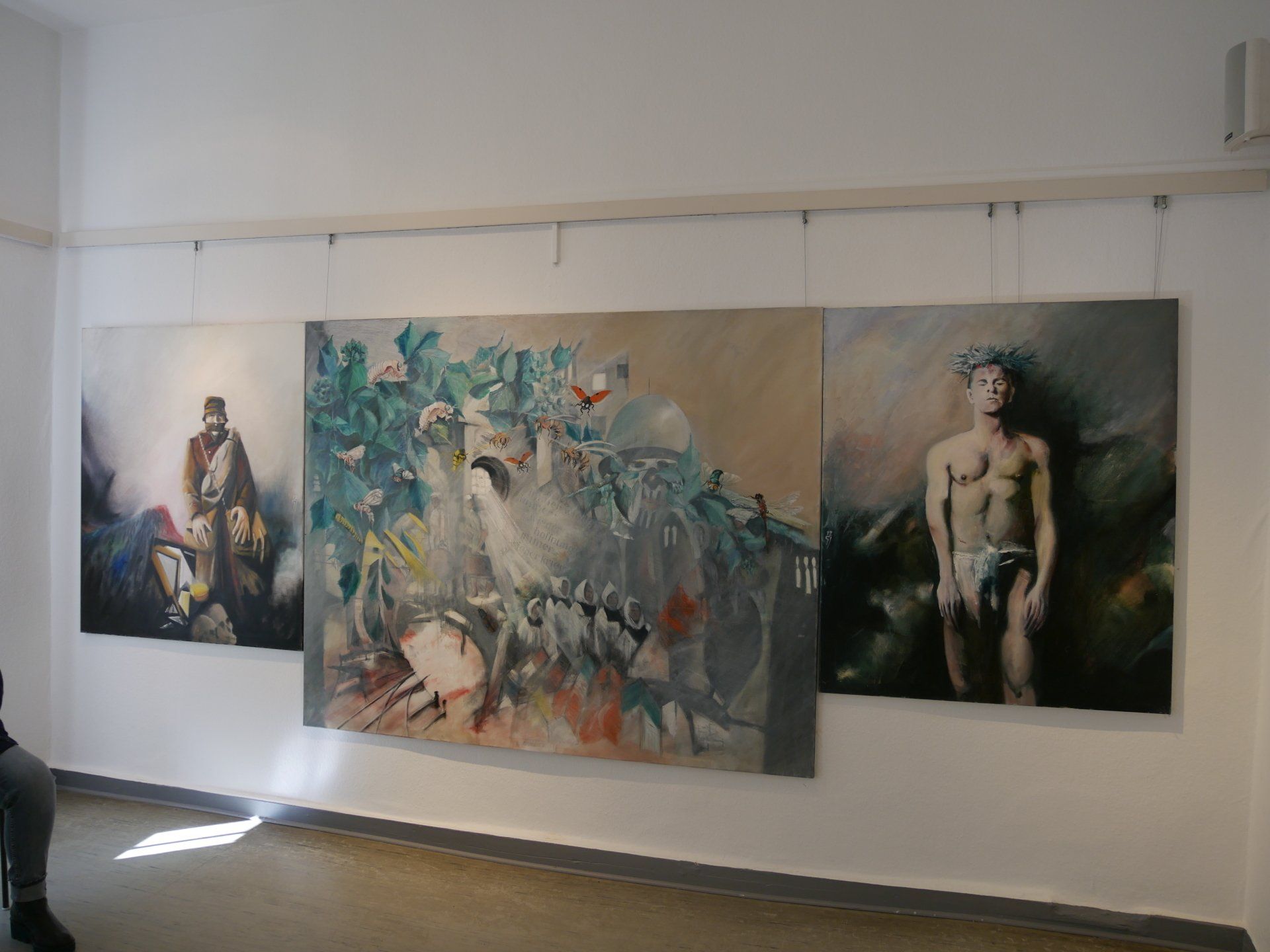 Drei großformatige figurative Gemälde hängen an einer weißen Galeriewand und werden durch Schienenbeleuchtung ergänzt.