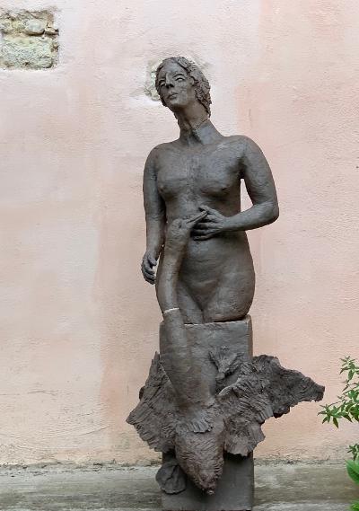 Bronzene Frauenstatue mit langem Hals und drapiertem Unterkörper, die vor einer hellen Wand steht.